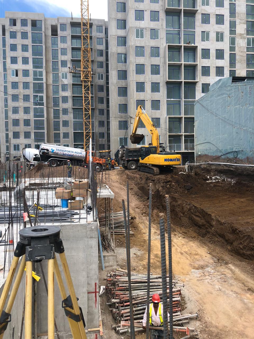 Torre de condominio en construcción con excavadora y estación total en primer plano